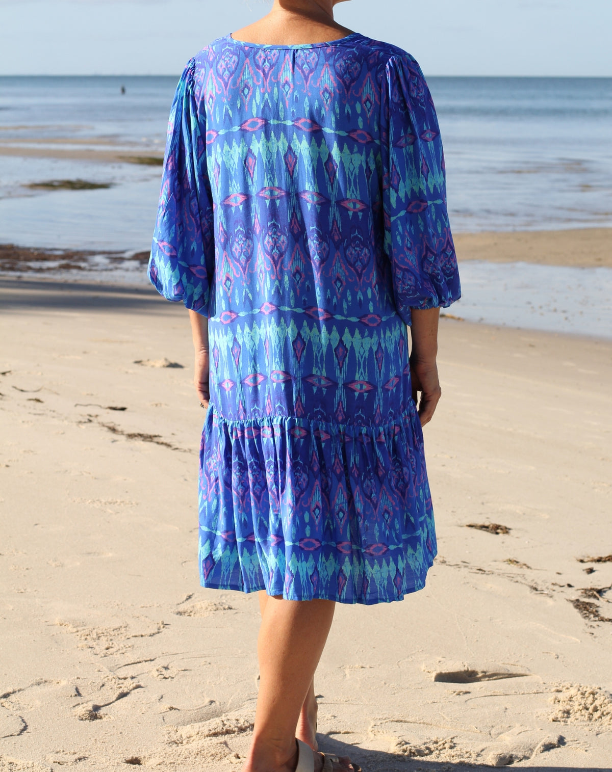 A woman stands on a sandy beach wearing a tiered boho midi dress with a blue, purple, and teal geometric pattern. The photo is taken from behind, showing the back of the dress with its three-quarter sleeves and ruffled hem.