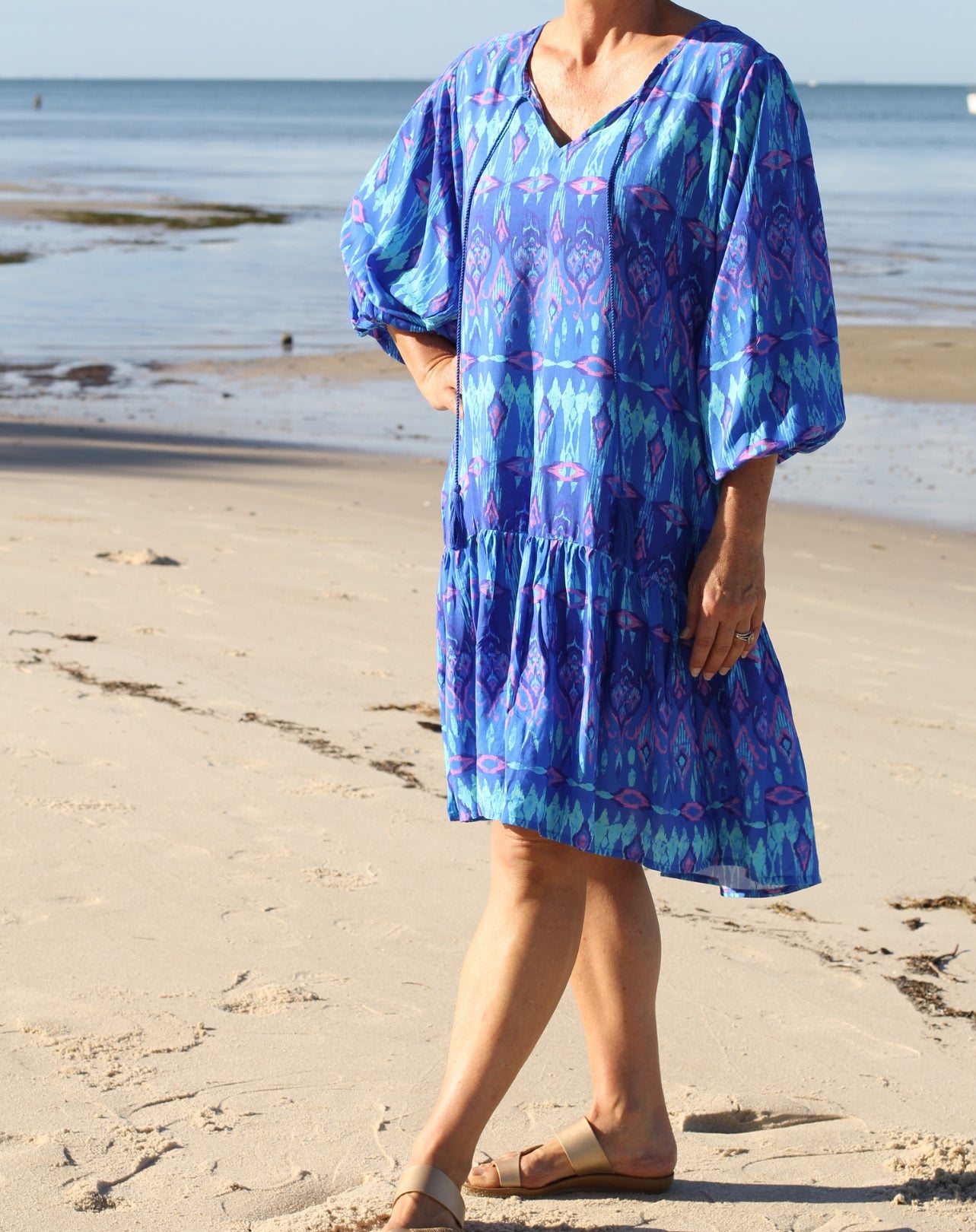 A midsized woman wearing a blue and purple ikat patterned tiered boho tunic dress with puff sleeves, standing on a sandy beach with the ocean in the background.