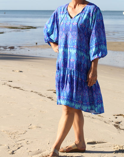 A midsized woman wearing a blue and purple ikat patterned tiered boho tunic dress with puff sleeves, standing on a sandy beach with the ocean in the background.