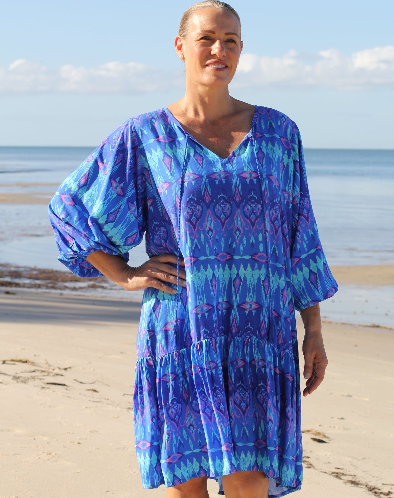 A woman wearing a blue and purple patterned bohemian-style tiered dress stands on a sandy beach with the ocean and sky in the background.