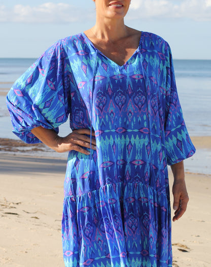 A woman wearing a loose, tiered bohemian-style dress with a blue, purple, and teal geo ikat pattern, standing on a sandy beach with the ocean in the background.