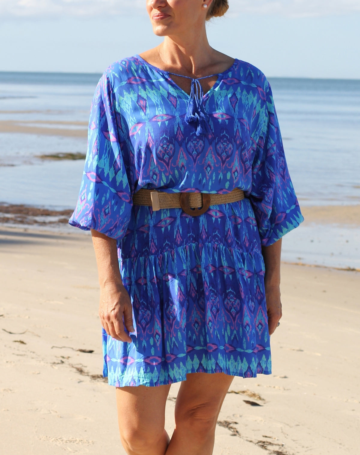 A woman standing on a sandy beach wearing a blue and purple ikat patterned kaftan-style dress with a tan belt.