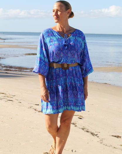 A woman stands on a sandy beach wearing a loose, short ikat blue patterned boho dress with three-quarter sleeves and a brown belt, with the ocean and sky in the background.