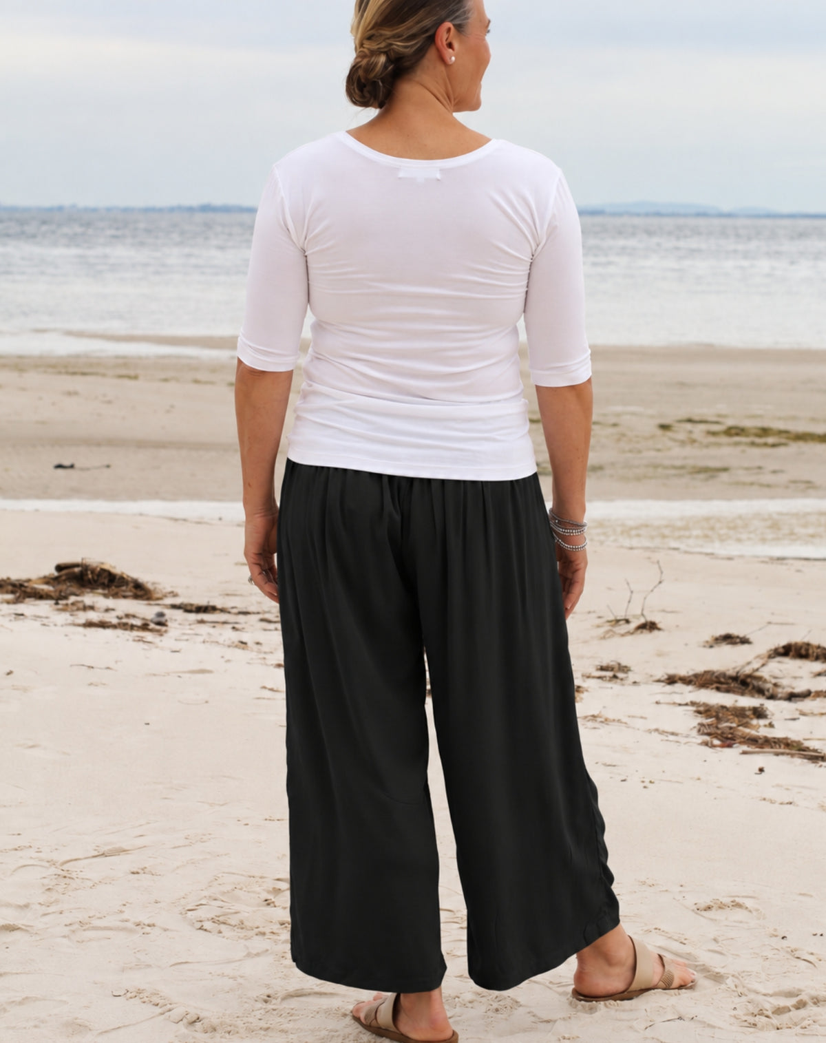 A woman standing on a sandy beach facing the ocean, wearing a white three-quarter sleeve top and loose black wide-leg pants.