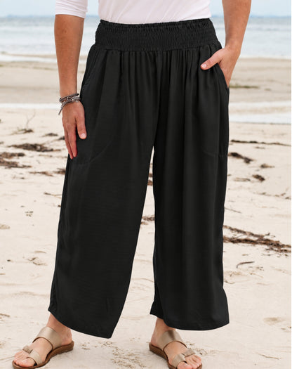 Black wide-leg cropped pants with an elastic waistband and side pockets, shown on a model standing on a sandy beach.