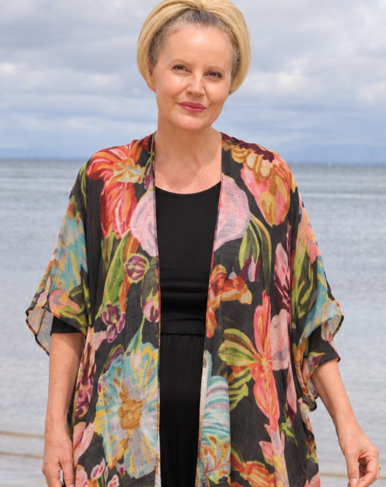 A woman standing outdoors by the water wearing a black top and a flowing open-front floral kimono cardigan with large multicolored flowers in pink, orange, green, and blue.