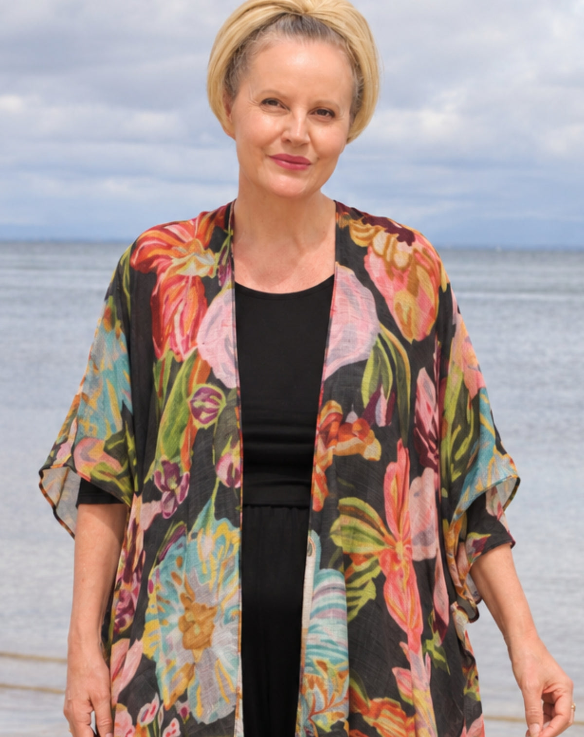 A woman standing outdoors by the water wearing a black top and a flowing open-front floral kimono cardigan with large multicolored flowers in pink, orange, green, and blue.