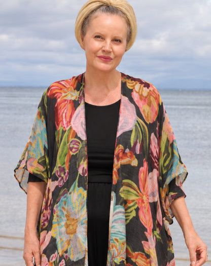 A woman standing outdoors by the water wearing a black top and a flowing open-front floral kimono cardigan with large multicolored flowers in pink, orange, green, and blue.