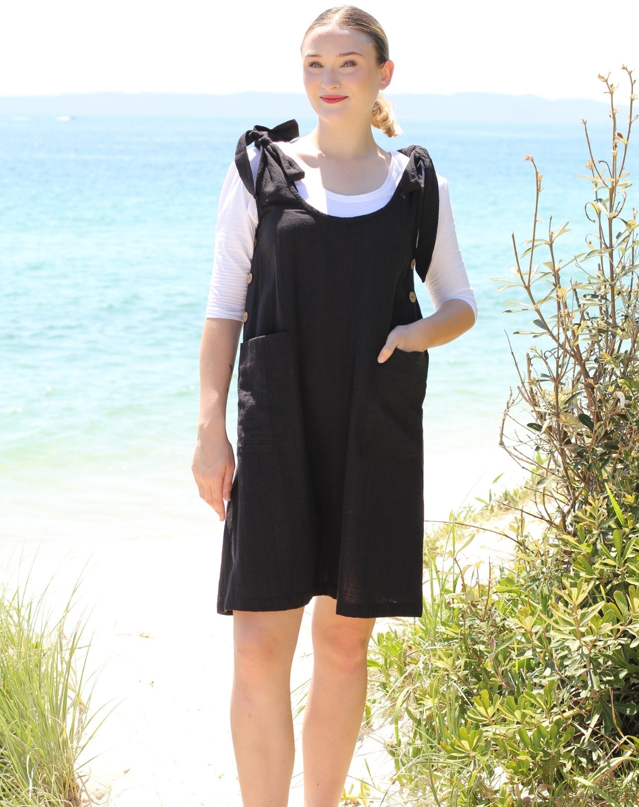 A size 8 woman standing on a beach wearing a black smock-style dress with a white t-shirt underneath. The background shows the blue ocean and sandy shore.