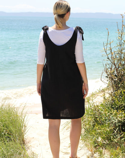 A woman stands on a sandy path by the ocean, viewed from behind, wearing a black sleeveless pinafore-style dress over a white three-quarter-sleeve top.