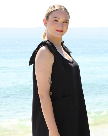 A woman with blonde hair tied back stands on a beach wearing a sleeveless black tie strap smock dress with a bow detail at the shoulders and a front pocket. The background shows the blue ocean and a bright sky.