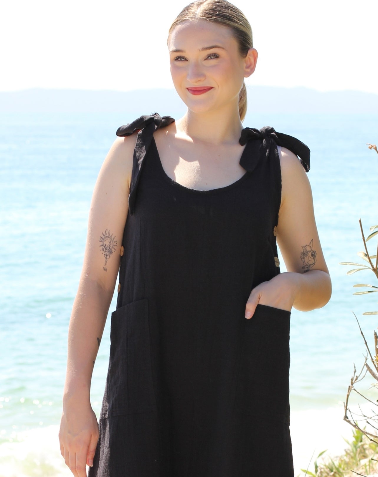 A woman wearing a black sleeveless smock dress with shoulder tie details and front pockets, standing outdoors with a bright blue ocean and sky in the background.