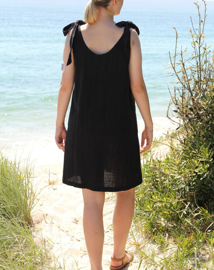 A woman viewed from behind wearing a sleeveless black dress with tie shoulder straps, standing on a sandy beach with ocean and greenery in the background.