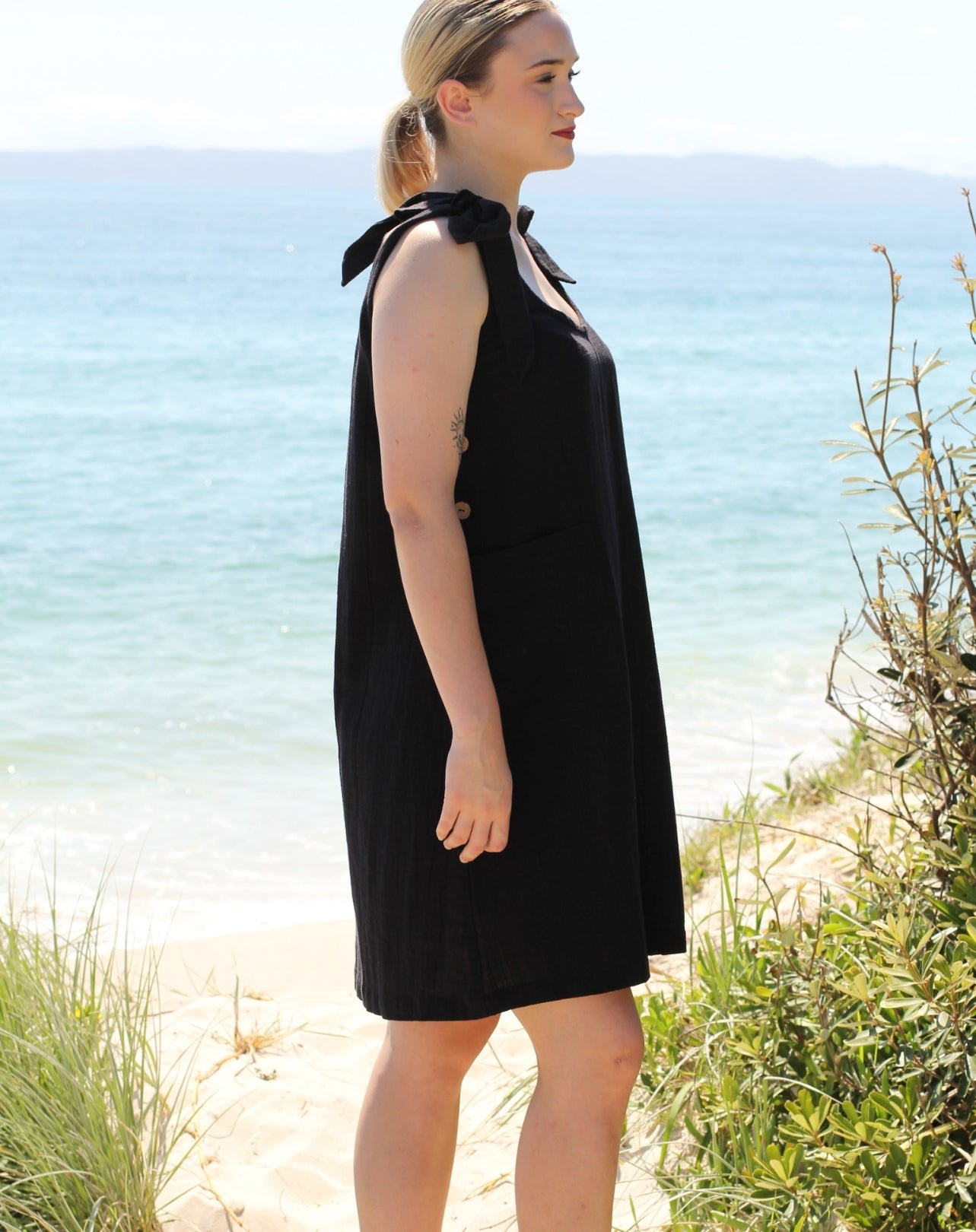 A woman wearing a sleeveless black smock dress with a tie-neck detail stands on a sandy beach with the ocean in the background.