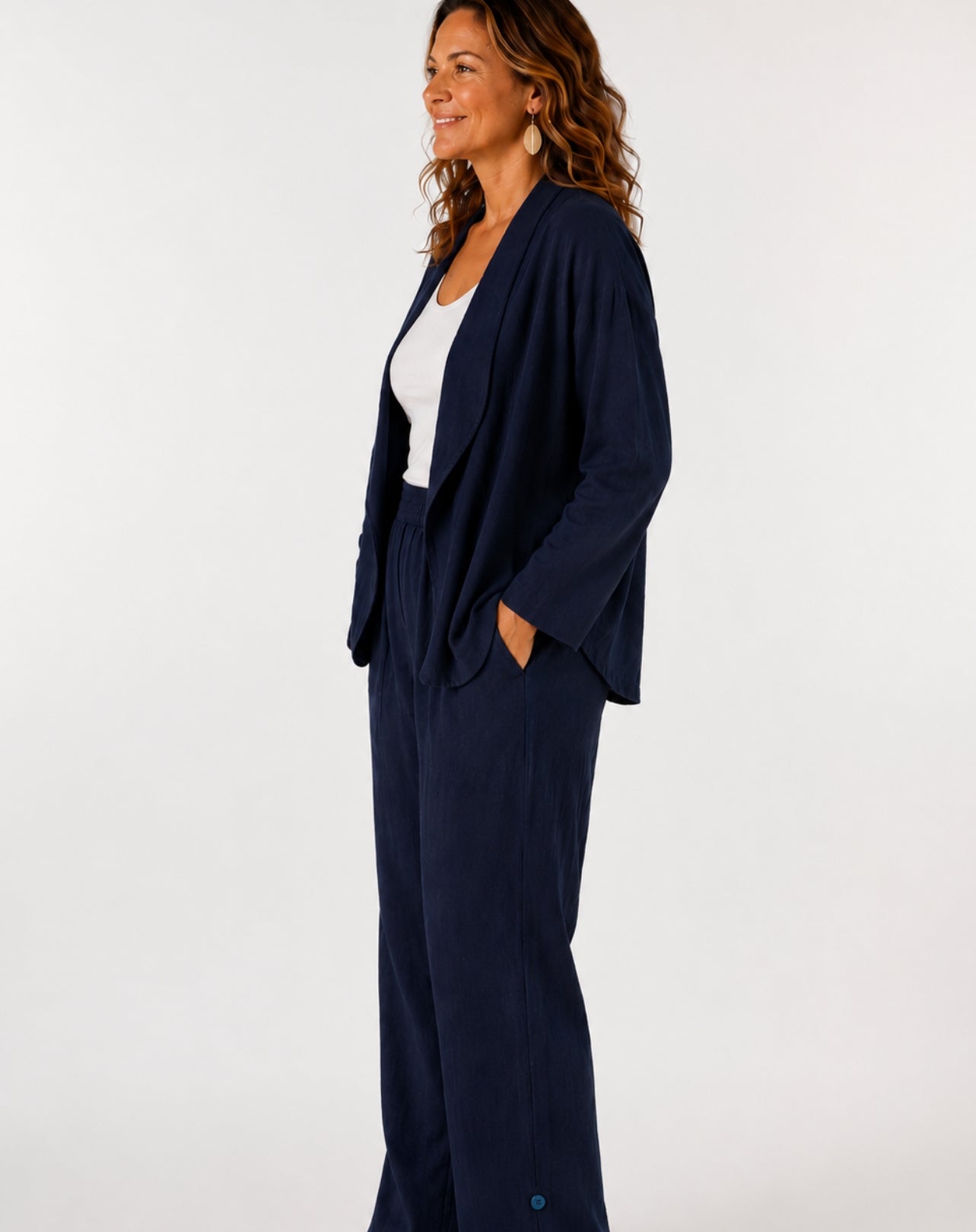 A woman wearing a navy blue two-piece linen look pant and blazer suit with a white top, standing in profile against a plain white studio background.