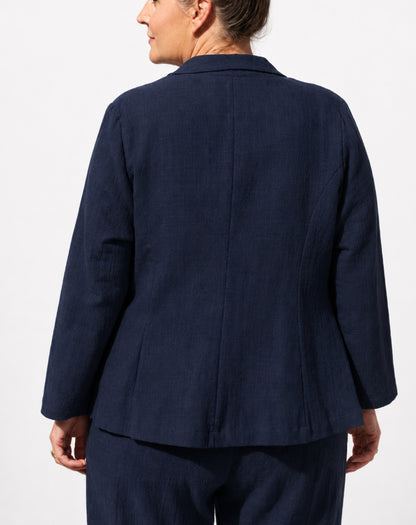A woman shown from the back wearing a dark navy blue tailored cotton linen jacket and matching trousers against a plain light background.
