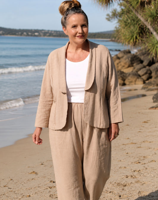 A woman standing on a sandy beach wearing a natural linen-blend two-piece set consisting of a draped open-front blazer jacket and matching straight-leg trousers, styled over a white scoop top.