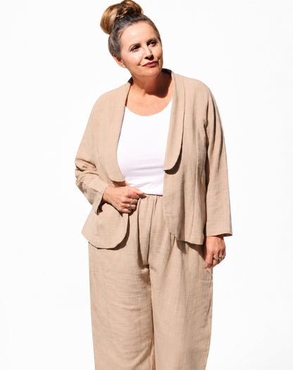 A woman modelling a natural cotton linen-blend two-piece set consisting of a draped open-front jacket and matching wide-leg trousers, worn over a white tank top, against a plain white background.