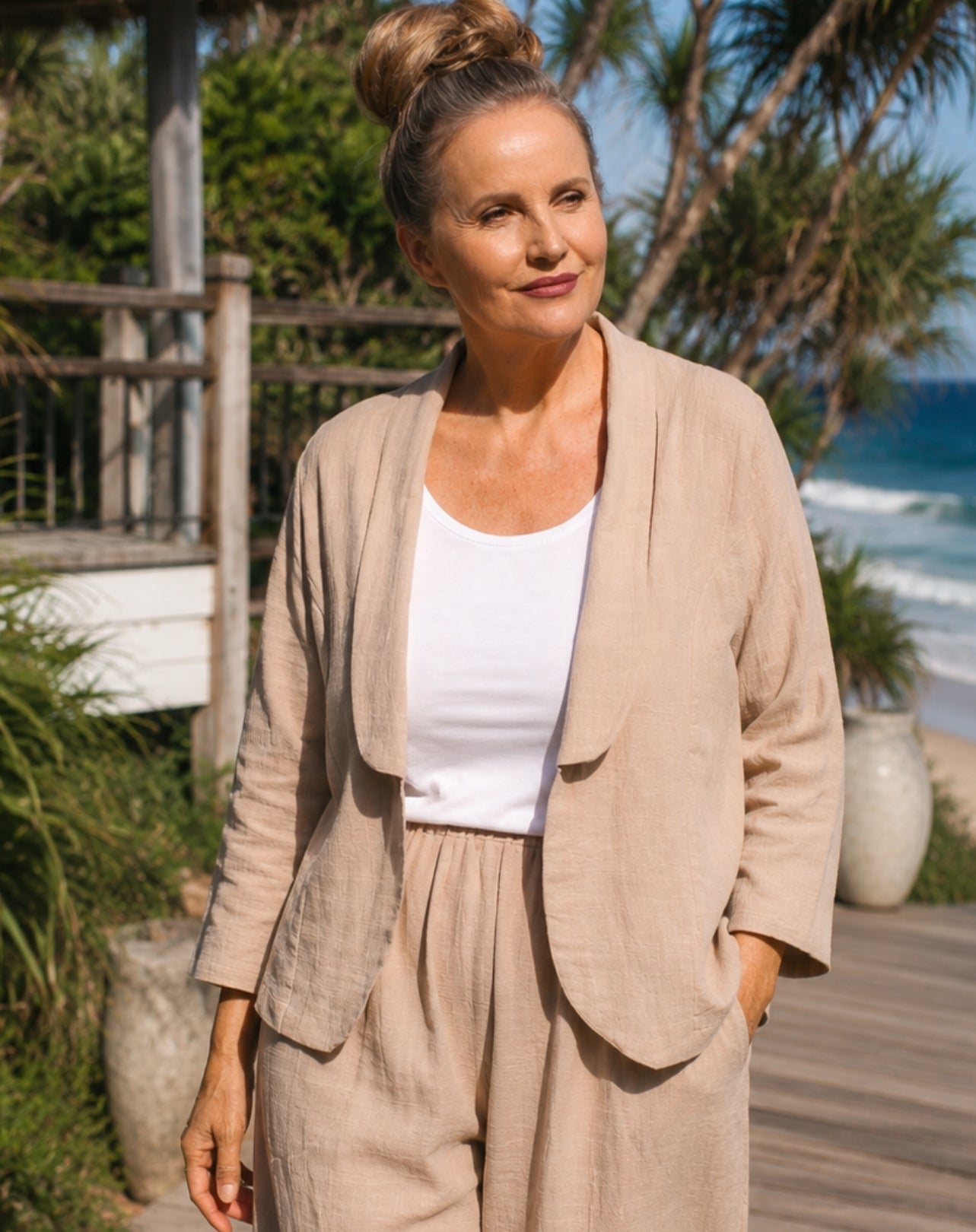 A woman wearing a beige linen-blend jacket and matching trousers over a white tank top, standing on a wooden boardwalk with a beach and palm trees in the background.