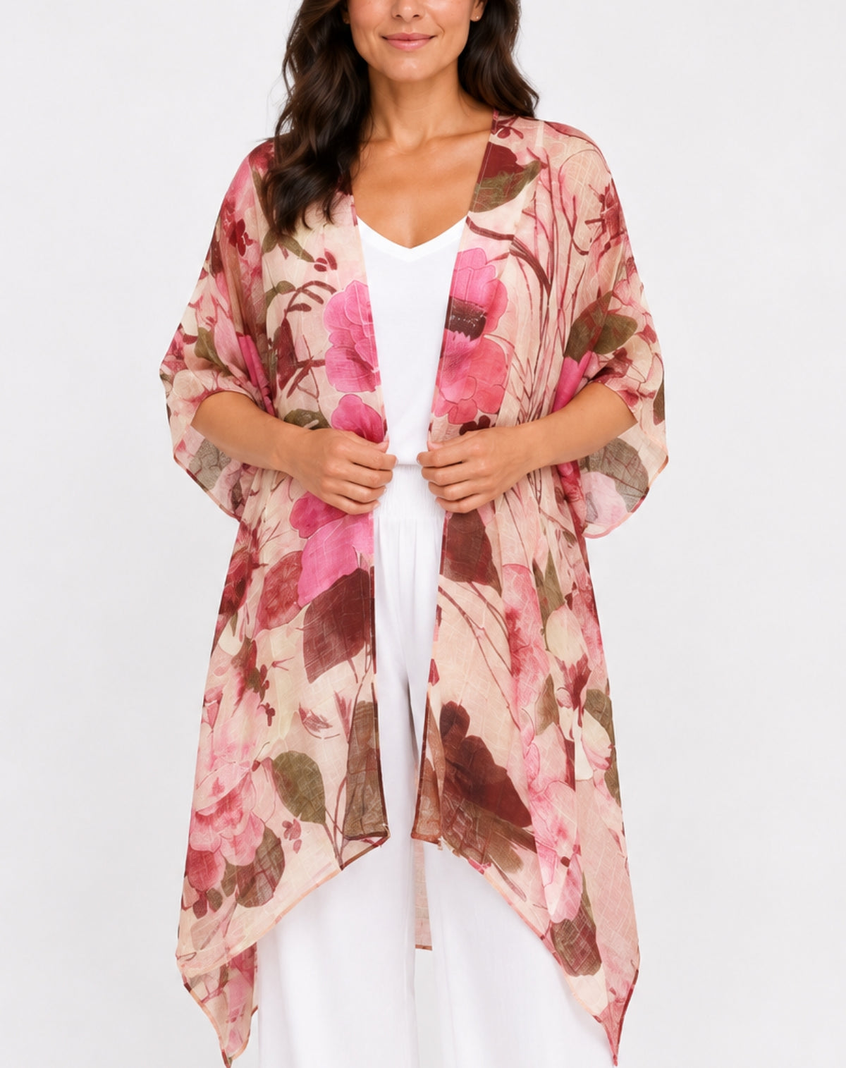 A woman wearing a lightweight floral kimono cardigan in pink, brown, and beige tones over a white top and white pants, photographed against a plain light gray studio background.
