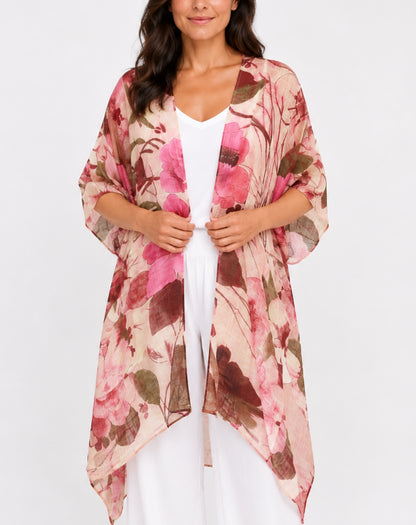 A woman wearing a lightweight floral kimono cardigan in pink, brown, and beige tones over a white top and white pants, photographed against a plain light gray studio background.