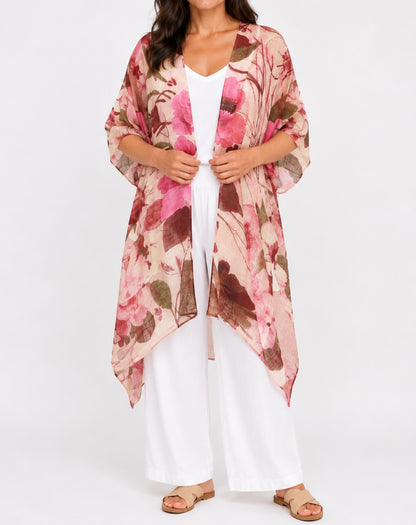 A woman models a lightweight floral kimono cardigan with pink, brown, and beige watercolor-style flowers over a white jumpsuit, set against a plain white studio background.
