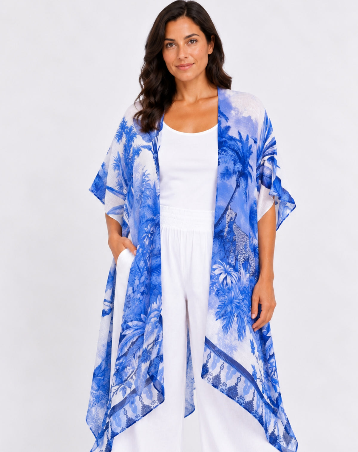 A woman modeling a lightweight open-front kimono-style cover-up with a blue and white cheetah and  tropical palm tree print, worn over a white tank top and white pants against a light gray studio background.