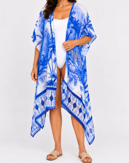 A woman modeling a lightweight blue and white tropical print kimono cover-up over a white swimsuit, paired with tan slide sandals, against a plain white studio background.