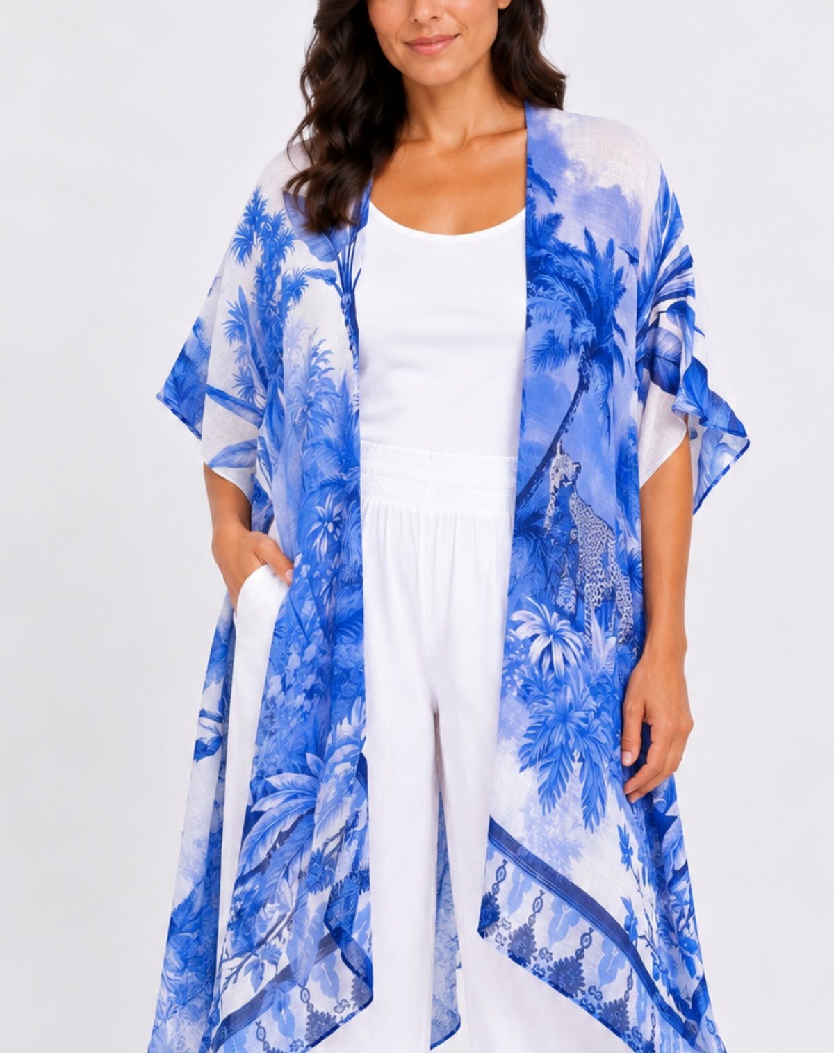 A woman wearing a blue and white tropical palm tree print kimono cardigan over a white tank top and white pants, photographed against a light gray studio background.