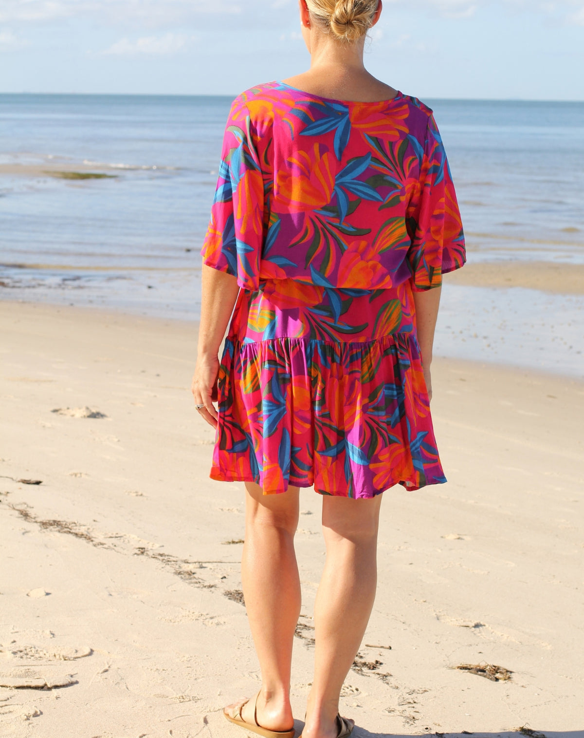 A woman standing on a sandy beach with the ocean in the background, wearing a short-sleeve boho flutter dress with a vibrant tropical floral print in pink, orange, purple, and blue.