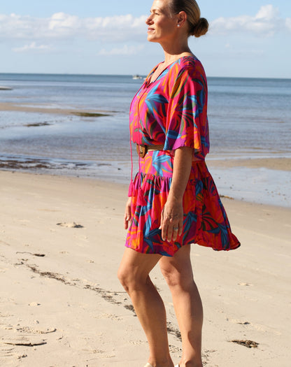 A woman wearing a vibrant tropical print dress stands on a sandy beach with the ocean and sky in the background.