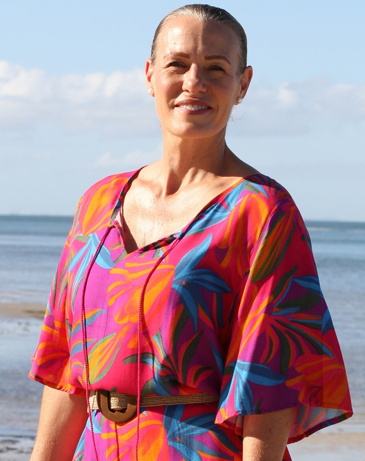 A woman standing on a beach wearing a vibrant, short-sleeved V-neck dress with a bold tropical floral pattern in pink, orange, blue, and purple, accessorized with a brown belt.