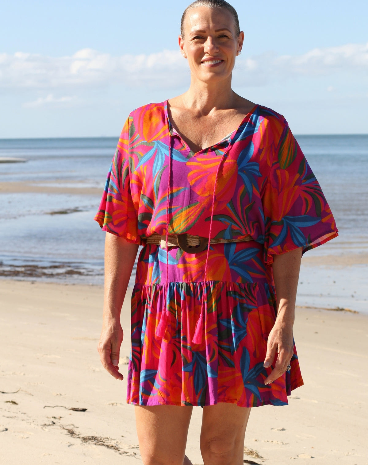 A woman standing on a sandy beach wearing a vibrant, multicolored tropical print kaftan dress with a tie neckline and a matching belt.