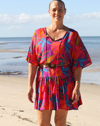A woman standing on a sandy beach wearing a vibrant, multicolored tropical print kaftan dress with a tie neckline and a matching belt.