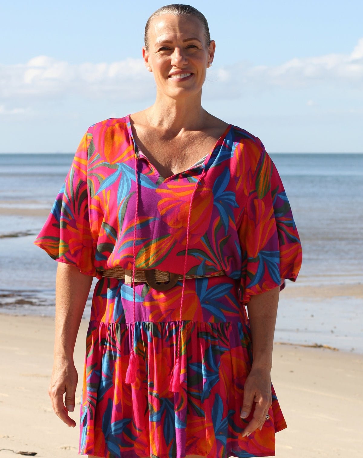 A woman standing on a sandy beach wearing a vibrant tropical floral dress with a brown belt. The dress features a bold pattern of pink, orange, blue, and red with a V-neckline and short puff sleeves.