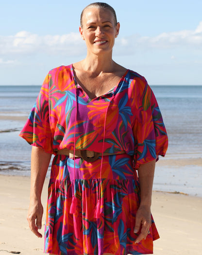 A woman standing on a sandy beach wearing a vibrant tropical floral dress with a brown belt. The dress features a bold pattern of pink, orange, blue, and red with a V-neckline and short puff sleeves.
