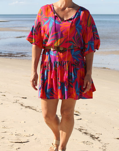A woman wearing a vibrant tropical print mini dress walks on a sandy beach with the ocean in the background. The dress features a bold pattern of pink, orange, blue, and green leaves with a matching belt.