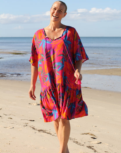 A woman smiling on a sandy beach wearing a vibrant, short-sleeved flutter dress with a tropical floral pattern in pink, orange, purple, and blue. She is holding a small pink tassel bag.