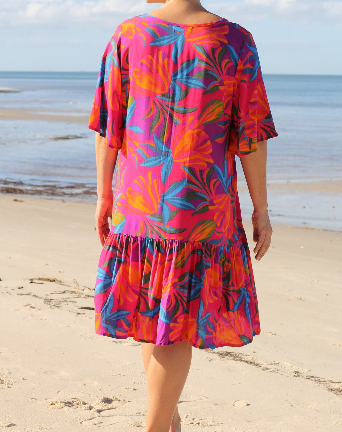 A woman standing on a sandy beach facing away from the camera, wearing a vibrant pink tiered dress with a colorful tropical floral pattern in orange, blue, and purple.