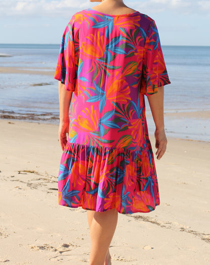 A woman standing on a sandy beach facing away from the camera, wearing a vibrant pink tiered dress with a colorful tropical floral pattern in orange, blue, and purple.