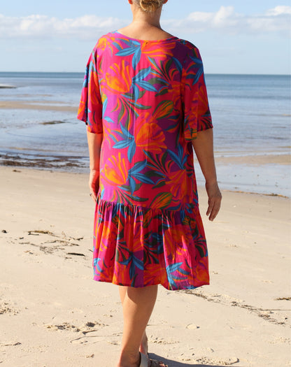 A woman viewed from behind wearing a short-sleeve tiered dress with a vibrant tropical floral print in pink, orange, and blue, standing on a sandy beach with the ocean in the background.