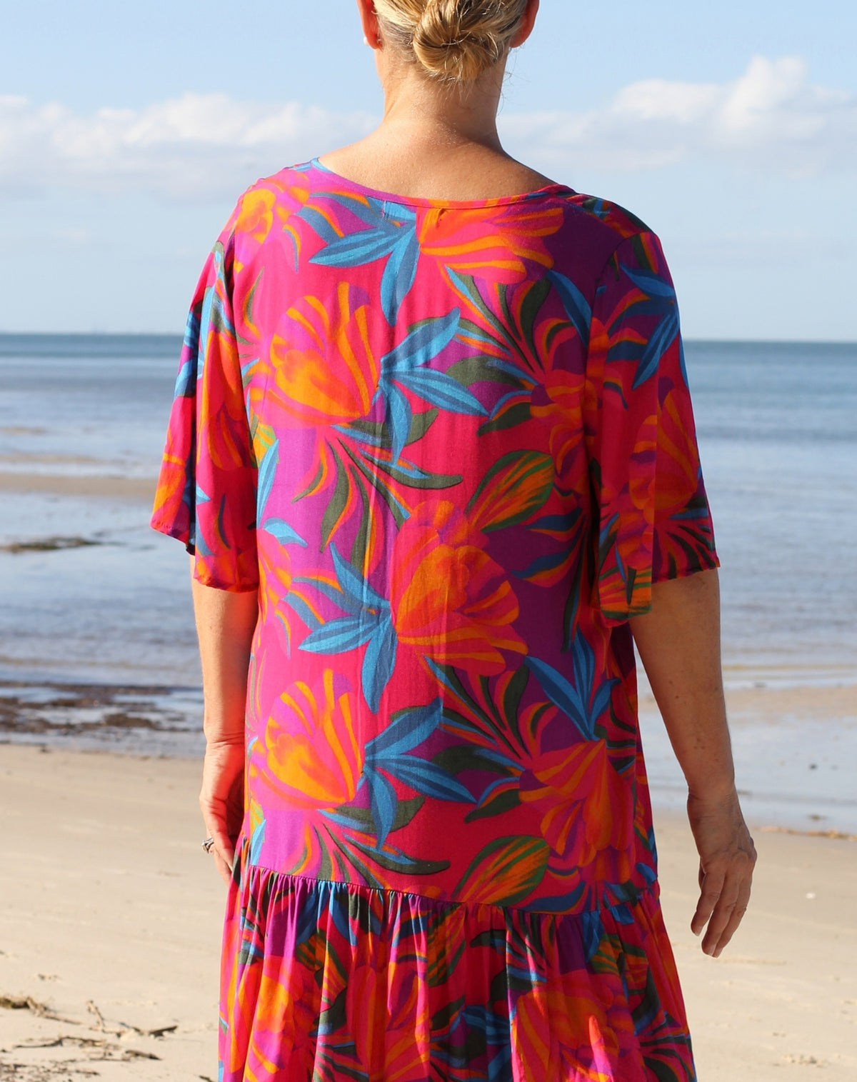 A woman standing on a sandy beach with the ocean and sky in the background, wearing a short-sleeve flutter dress with a bold tropical floral print in pink, orange, blue, and purple.