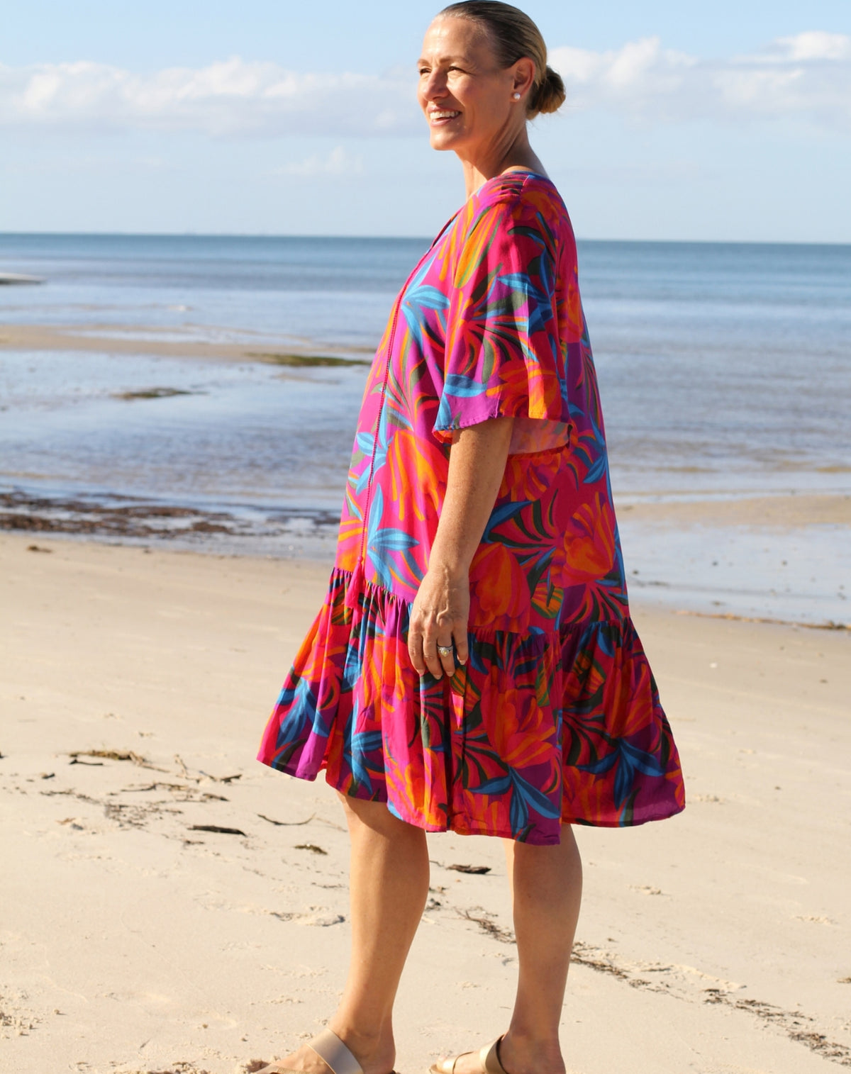 A woman wearing a vibrant tropical print dress stands on a sandy beach with the ocean and sky in the background. The dress features a bold pattern of pink, orange, red, and blue leaves.