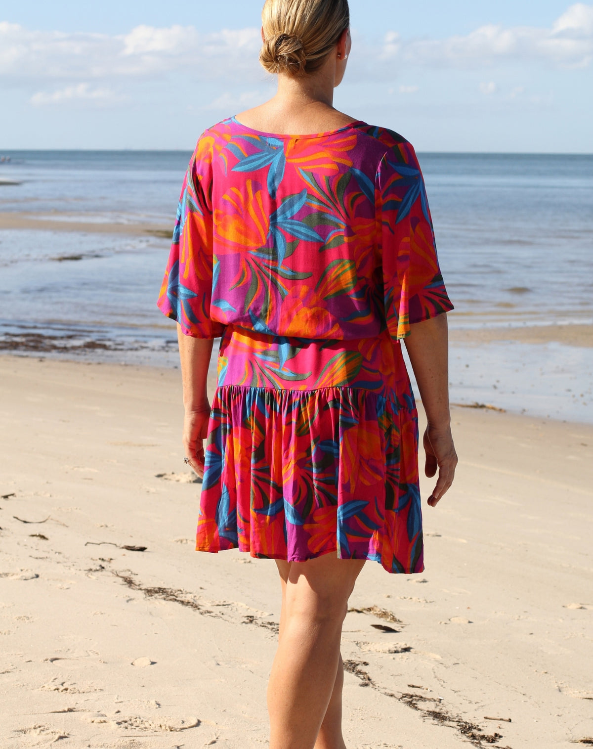 A woman viewed from behind wearing a vibrant, multicolored tropical print boho flutter dress while standing on a sandy beach with the ocean in the background.