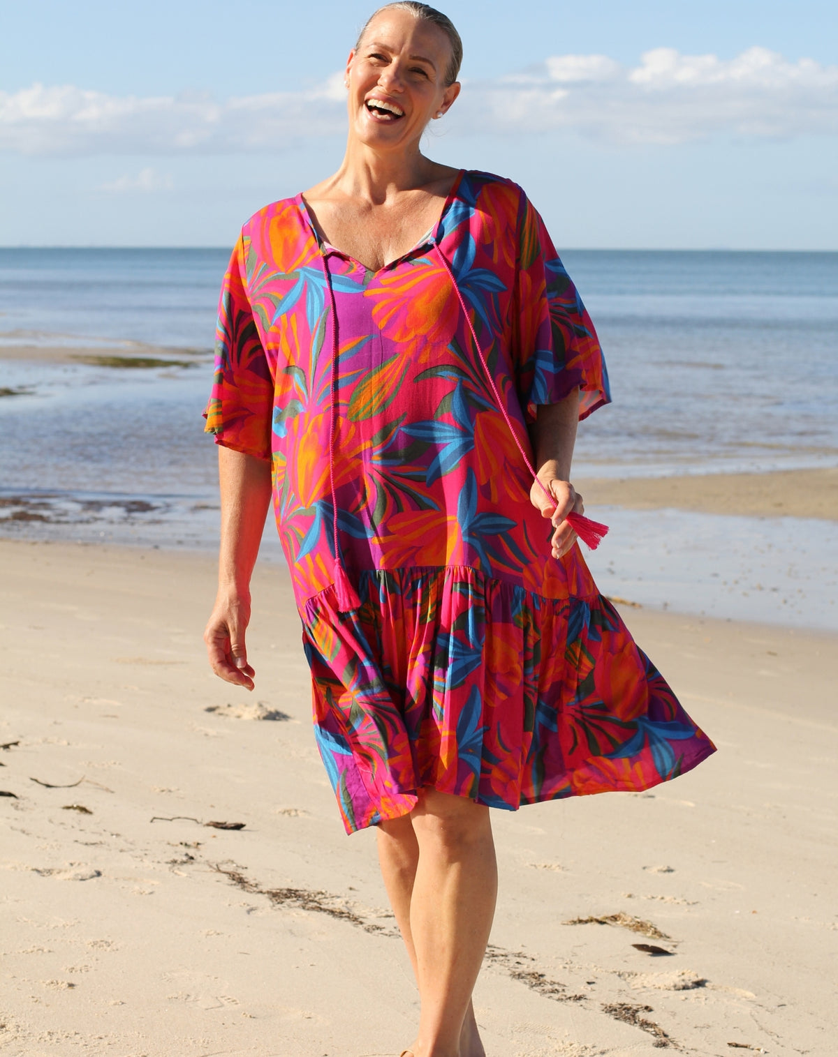 A woman smiling and walking on a sandy beach wearing a vibrant, tiered boho flutter dress with a bold abstract floral pattern in red, pink, orange, and blue.