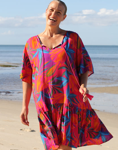 A woman smiling on a sandy beach wearing a tiered flutter short-sleeve dress with a vibrant tropical floral pattern in pink, orange, blue, and purple.