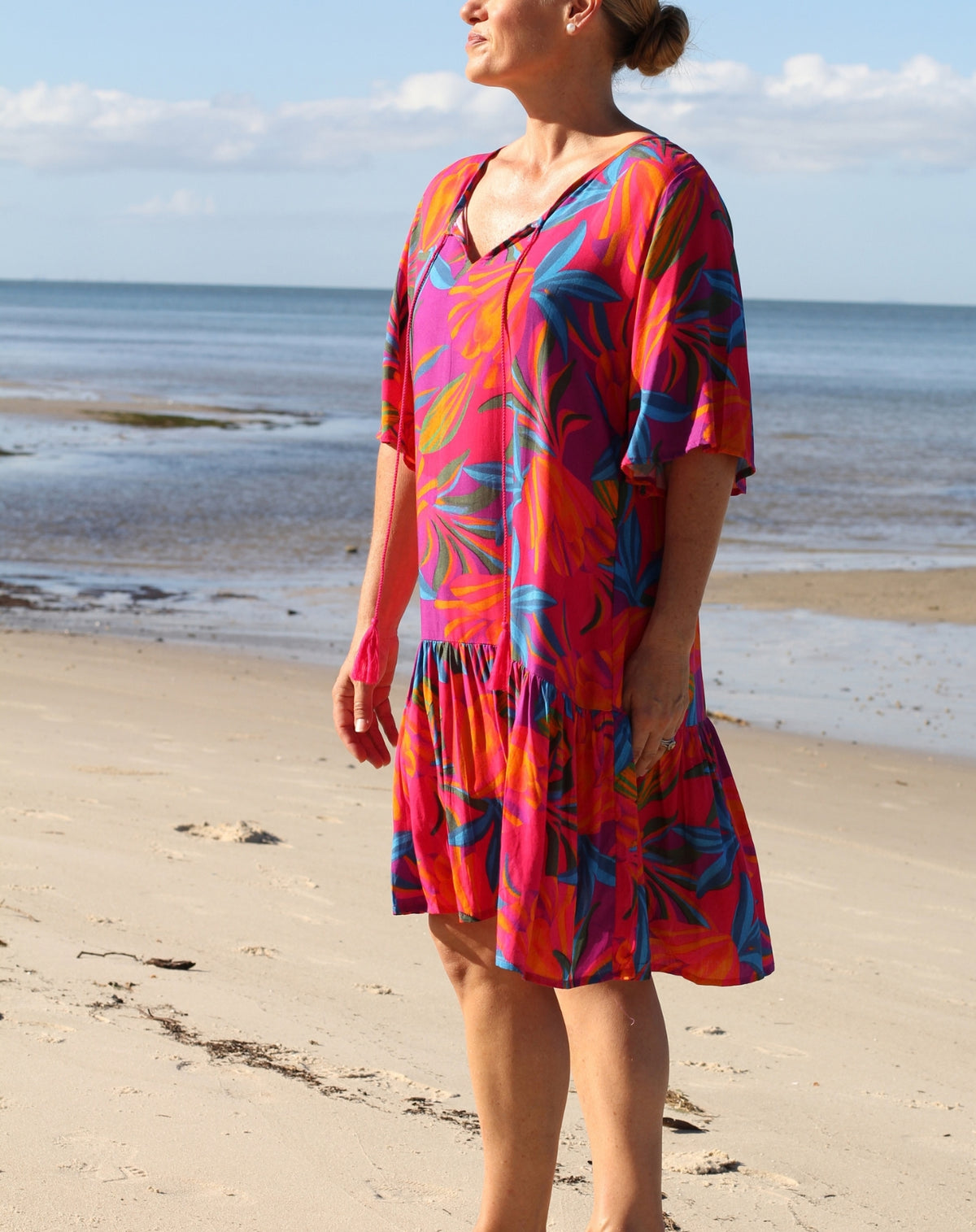 A woman wearing a vibrant tropical print tunic dress stands on a sandy beach with the ocean and sky in the background. The dress features a bold pattern of pink, orange, blue, and yellow leaves.