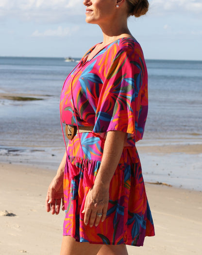 A woman wearing a vibrant, multicolored tropical print dress stands on a sandy beach with the ocean and sky in the background.