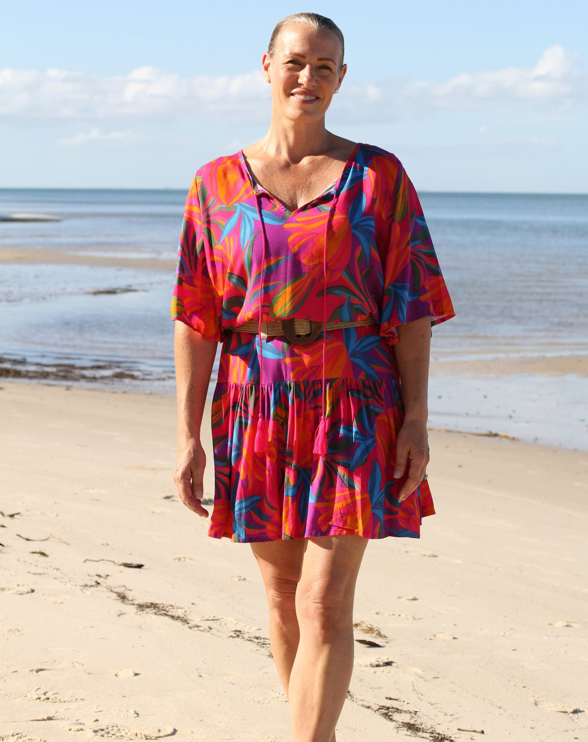 A woman walking on a sandy beach wearing a short, tiered boho flutter dress with a vibrant tropical floral pattern in red, orange, pink, and blue, accessorized with a brown belt.