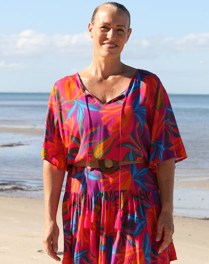 A woman stands on a sandy beach wearing a bright tropical floral dress with pink, orange, blue, and purple tones, a brown belt, and a ring, with the ocean and sky in the background.